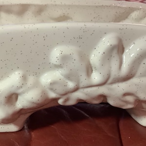 May include: A white ceramic planter with a speckled finish and a raised leaf design. The rectangular planter has a curved shape and sits on small feet. The leaf design is a prominent feature, adding a touch of nature to the piece.