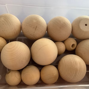 May include: A collection of unfinished wooden spheres of varying sizes. The spheres are light brown in color and appear to be made of natural wood. Some spheres have a hole drilled through them. The spheres are in a clear plastic container.