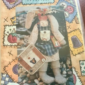 May include: A handmade bunny doll wearing a floral crown, apron, and holding a book. The doll has long ears and is dressed in a cream-colored dress with a blue plaid apron. The doll is holding a small book and a square sign that says "FLOWERS".