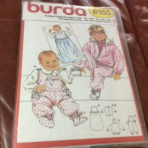 May include: A Burda 6155 sewing pattern for babies and toddlers. The pattern includes illustrations of a baby in a dress, a baby in overalls, and a toddler in a pink tracksuit. The pattern sizes range from 3 months to 3 years.