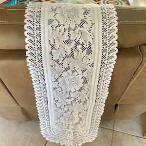 May include: A white lace table runner with a floral pattern. The runner is rectangular and features intricate detailing along the edges and center. The delicate design adds a touch of elegance to any table setting.