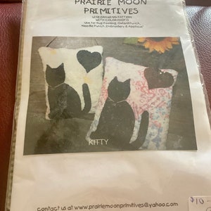 May include: A package for a craft pattern from Prairie Moon Primitives. The pattern features two pillows with black cat silhouettes and heart accents. The word "KITTY" is printed below the image. The pattern is for rug hooking, Oxford punch, needle punch, embroidery, and appliqué.