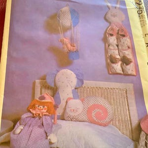 May include: A vintage McCall's pattern featuring nursery decor. The image shows a doll, an elephant pillow, a snail pillow, a hot air balloon mobile, and a bunny-shaped shoe organizer. The color palette includes soft pastels like pink, blue, and lavender.