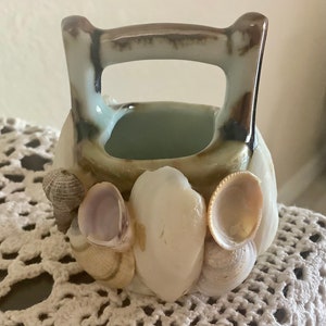 May include: A small, ceramic basket with a handle, adorned with various seashells. The basket is primarily off-white with brown accents on the handle. The seashells are in shades of white, tan, and light purple.