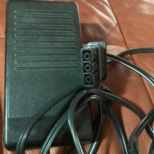 May include: Black sewing machine foot pedal with a textured surface and a three-prong plug. The pedal is connected to a black power cord. The image is of a sewing machine accessory.