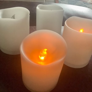 May include: Four white, battery-operated LED candles with a soft, frosted appearance. Two candles have a flickering amber light, while the other two are unlit. The candles are arranged on a dark brown surface.