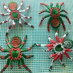May include: Four handmade spider decorations. Each spider is crafted with red, green, and white yarn, featuring detailed patterns on the body and legs. The spiders are arranged on a green grid background, showcasing their intricate designs.