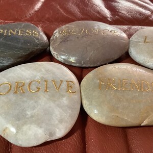 May include: Five smooth, oval stones with gold lettering. The stones display the words "HAPPINESS", "REJOICE", "LOVE", "FORGIVE", and "FRIENDSHIP". The stones vary in color from gray to white and beige.