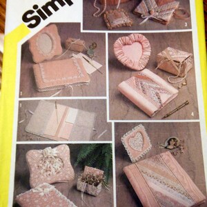May include: Simplicity sewing pattern featuring various craft projects. The image showcases several items, including a heart-shaped frame, a small box, and a book-shaped item, all in shades of pink and adorned with lace and bows. The pattern is by marjorie puckett.