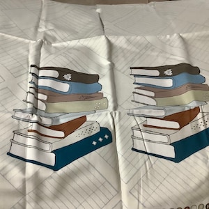 May include: White fabric with a repeating pattern of stacked books in various colors, including blue, brown, and gray. The books have a simple, illustrated style, and the background features a subtle grid design.