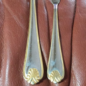 May include: Two pieces of silverware, a fork and a spoon, with a gold-colored trim and a decorative gold design at the base of the handle. The silverware is set against a brown leather surface.