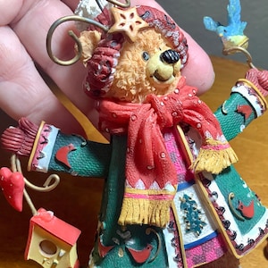 May include: A whimsical Christmas ornament featuring a bear character wearing a red scarf and green coat. The bear holds a blue bird and a branch with a red heart and birdhouse. The ornament has a star on its head and is decorated with festive details.