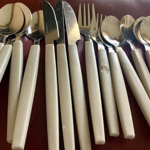 May include: A collection of stainless steel silverware with off-white handles. The set includes spoons, knives, and forks. The silverware is arranged on a red surface, showcasing a vintage or retro style.