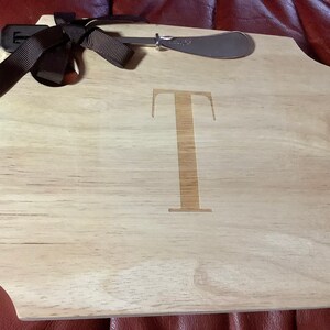 May include: Wooden serving board with a light brown finish, featuring a large letter "T" in the center. Includes a stainless steel spreader and a dark brown ribbon. Ideal for serving cheese and appetizers.