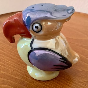 May include: A ceramic parrot-shaped salt shaker with a pearlescent finish. The bird features a red beak, blue and purple accents, and a yellow body. The top has several holes for dispensing salt. A decorative kitchen item.
