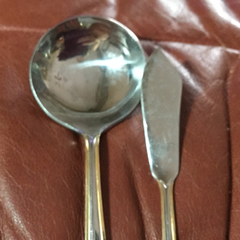 May include: A stainless steel ladle and butter knife set with gold-colored accents. The ladle has a round bowl and a long handle. The butter knife has a flat, angled blade. The set is displayed on a brown surface.