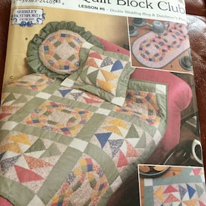 Pode incluir: Um padrão Simplicity Quilt Block Club para a Lição nº 6, Double Wedding Ring & Dutchman's Puzzle. A imagem mostra um edredom, uma almofada e uma almofada redonda com padrões de patchwork coloridos em tons de verde, azul, amarelo e vermelho.
