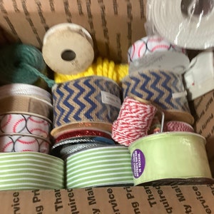 May include: A cardboard box filled with various spools of ribbon. The ribbons include baseball-themed, striped, and chevron patterns in colors like red, white, blue, and green. There is also a ball of green yarn and yellow rope.