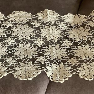 May include: Cream-colored, open-weave lace table runner. The runner features a repeating floral pattern with scalloped edges. The delicate design is visible against the dark background, highlighting the intricate craftsmanship. This decorative piece adds a touch of elegance to any table setting.
