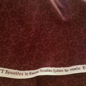May include: Dark brown fabric with a floral pattern. The fabric has a repeating design of stylized flowers in a darker shade of brown. A white strip with the text "KT Favorites by Kansas Troubles Quilters for moda" is visible.