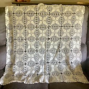 May include: Cream-colored, handmade crochet blanket with a repeating geometric pattern. The blanket is draped over a gray sofa, showcasing its intricate openwork design. The pattern features diamond and star-like shapes, creating a textured, decorative piece.