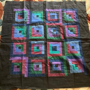 May include: A dark brown quilt with a log cabin pattern. The quilt features sixteen squares, each with a central dark square surrounded by concentric rectangles in shades of blue, green, red, and purple. The quilt is made of fabric.