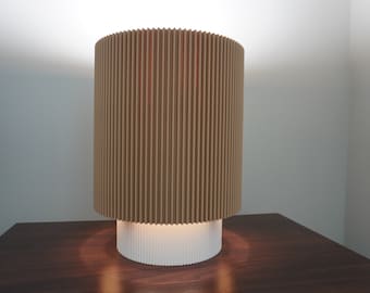 Op de afbeelding: Een moderne tafellamp met een bruine geplooide stoffen kap en een witte voet. De lamp is aan en verlicht het oppervlak eronder.