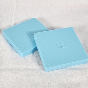 May include: Two light blue square boxes with a textured surface. The boxes have the letters "UE" embossed on the top. The boxes are on a white, textured surface.
