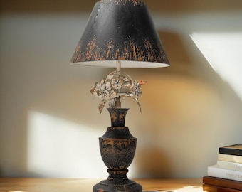 May include: A decorative table lamp with a black lampshade and a distressed, antique-style finish. The lamp features a floral accent and a black base with gold accents. The lamp is on a wooden surface.