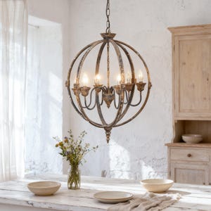 May include: A spherical, antique-style chandelier with six lights hangs above a rustic wooden table. The chandelier has a distressed, bronze finish and is suspended from a chain. The room features a white brick wall and a wooden cabinet.