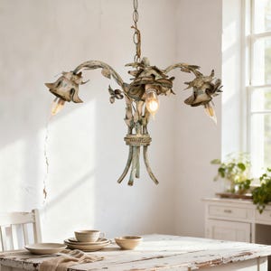 May include: A distressed, antique-style chandelier with three light fixtures. The metal frame has a floral design with a faded blue-green finish. The chandelier hangs above a rustic wooden table set with neutral-toned dishes.