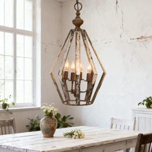 May include: A distressed, diamond-shaped chandelier with four candle-style lights hangs above a white wooden table. The metal frame has a weathered, off-white finish. A vase of flowers and a bouquet sit on the table, suggesting a dining room setting.