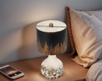May include: A table lamp with a black and gold lampshade and a white, textured base. The lamp is on a wooden bedside table next to a smartphone and a bed with a pillow.