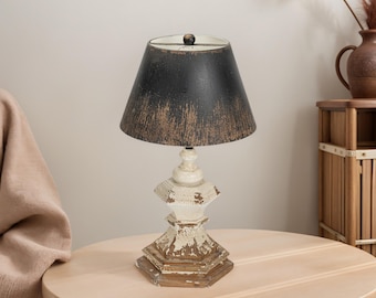 May include: A vintage-style table lamp featuring a black lampshade and a distressed white base. The lamp is placed on a round wooden table. The lampshade has a textured, aged look. The lamp's base has a unique, ornate design, adding to its antique appeal.