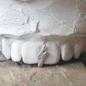 Puede incluir: Un molde de yeso blanco de un conjunto de dientes con un amuleto de rayo plateado entre los dientes delanteros.