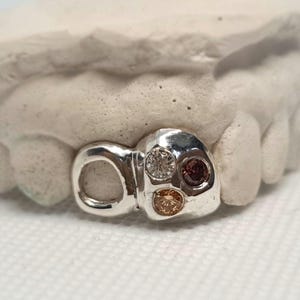 May include: A silver tooth grill with three colourful gemstones. The grill is designed to fit over a tooth and has a rounded shape. The gemstones are a clear diamond, a pink stone, and a yellow stone.