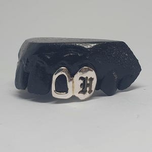 Puede incluir: Una pieza de grillz plateada que presenta dos fundas dentales, una con un diseño recortado y la otra grabada con la letra H en una fuente gótica, montada sobre un molde dental negro para mostrar el acabado metálico y el detalle.