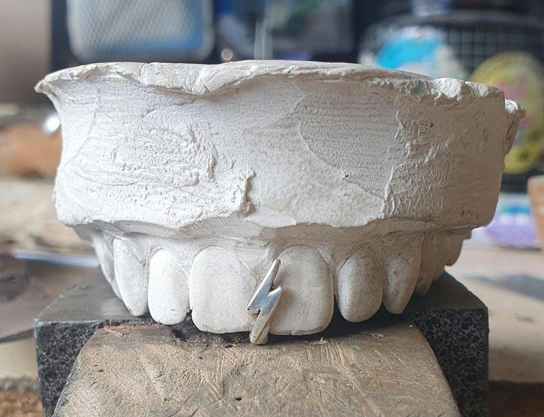 Puede incluir: Un molde de yeso blanco de un conjunto de dientes con un amuleto de rayo plateado entre los dientes delanteros.