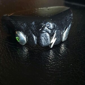 Puede incluir: Una parrilla negra y plateada con una piedra preciosa verde y un dise&ntilde;o de rayo.