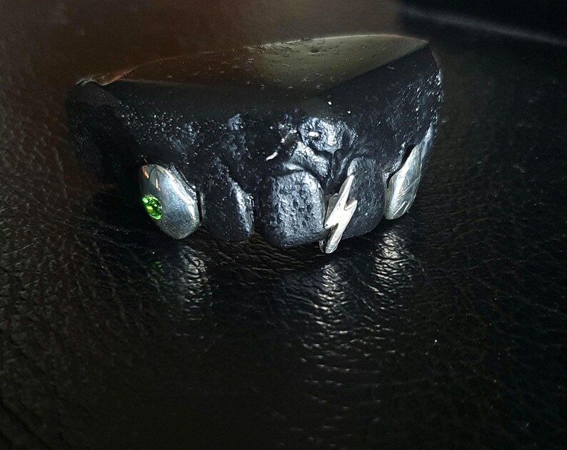 Puede incluir: Una parrilla negra y plateada con una piedra preciosa verde y un dise&ntilde;o de rayo.