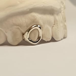 May include: A single silver-colored tooth grill with a smooth, polished finish, fitted onto a white dental mold. The grill features a circular opening in the center and is positioned over the front portion of the plaster dental model.