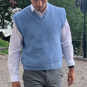 Pode incluir: Um colete de malha azul claro com decote em V. O colete tem um padrão texturizado e é usado sobre uma camisa branca de botões.