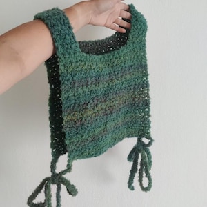 May include: A handmade, green crochet bag with a textured, fuzzy appearance. The bag has a square shape with a wide strap and two decorative ties at the bottom. The color is a mix of greens and blues.