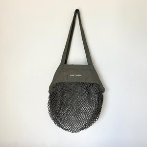 May include: An olive green tote bag featuring a solid fabric upper section with the text DON'T JUDGE printed in white, and a lower section made of a dark grey mesh net. The bag includes two long shoulder straps for carrying.