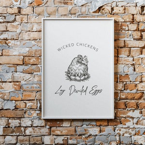 Pode incluir: Uma impressão emoldurada branca com uma ilustração em preto e branco de uma galinha a pôr ovos diabólicos. O texto diz "Wicked Chickens Lay Deviled Eggs".