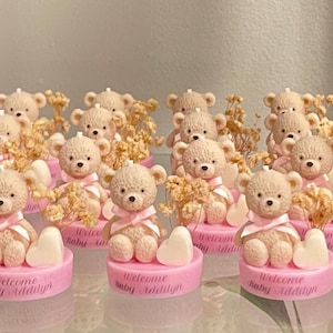 May include: A collection of teddy bear favors for a baby shower. Each bear is light brown with a pink ribbon and sits on a pink base with the text "Welcome Baby Addilyn". Includes dried flowers and a white heart.