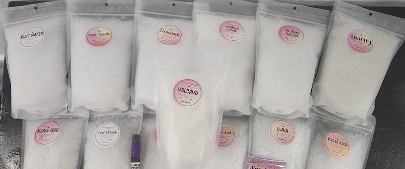 May include: A collection of 12 clear plastic bags filled with white bath salts. Each bag has a label with a different scent name, including "Drift Away", "Pink Sands", "Lemonade", "Volcano", "Lavender Lemonade", "Vanilla Bean", "Ocean Cider", "Coconut", "Calamity", "Liana", "White Lilac", and "Detoxing".