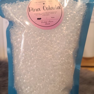 May include: A blue resealable bag filled with clear scented aroma beads. The label reads "The Freshie Shack" and "Pina Colada" with a warning to keep out of reach of children and pets. The label also states "Scented Aroma Beads" and "16 oz."
