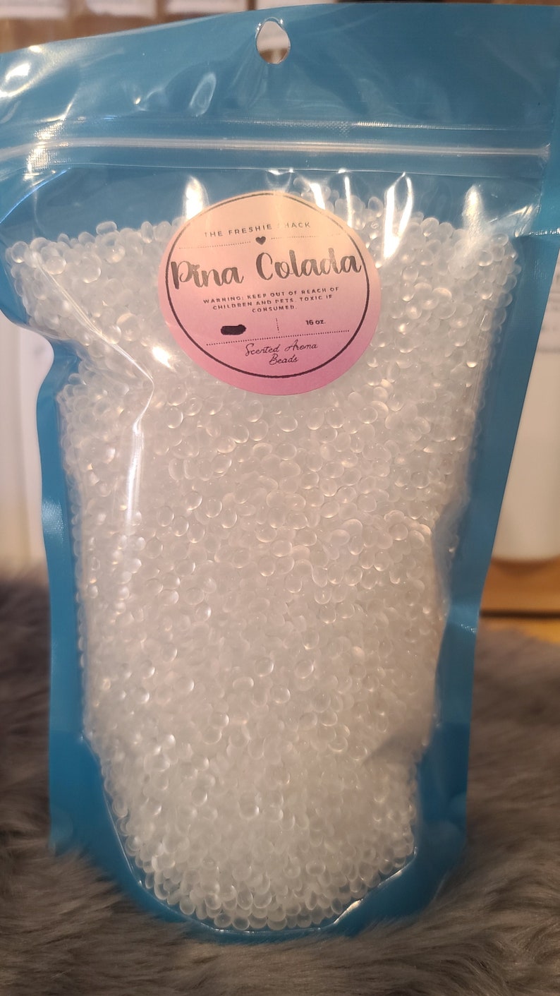 May include: A blue resealable bag filled with clear scented aroma beads. The label reads "The Freshie Shack" and "Pina Colada" with a warning to keep out of reach of children and pets. The label also states "Scented Aroma Beads" and "16 oz."