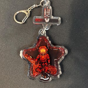 May include: A clear acrylic keychain featuring a red astronaut in a spacesuit on a red star-shaped background. The keychain has a silver keyring and a cross-shaped charm with a rocket design.